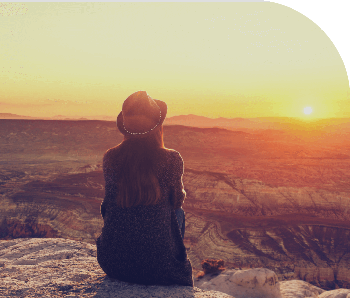 A woman sitting on top of a mountain looking at the sun.